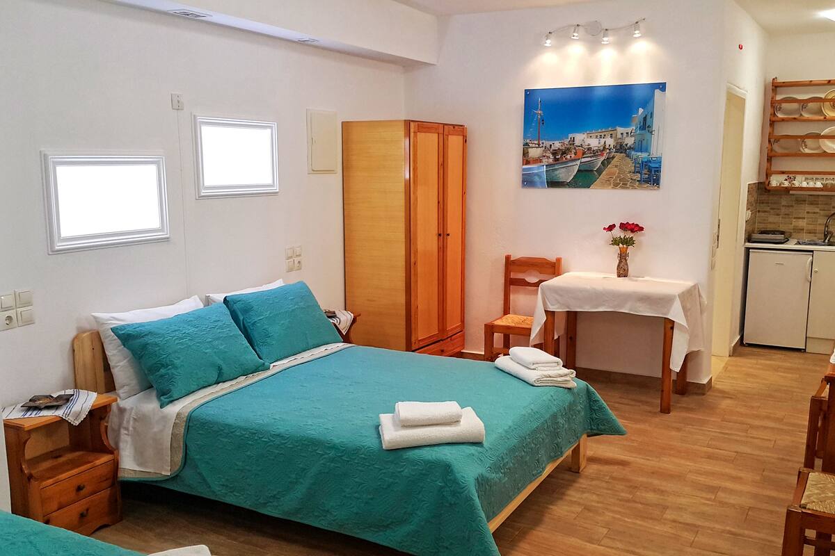 A bedroom with a bed, a wooden wardrobe, a small table with a vase, and a view of a coastal scene.