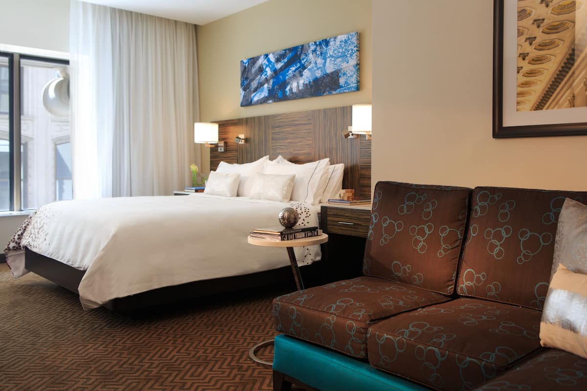 A hotel room with a large bed, a sofa, a small table with books, and a painting on the wall.