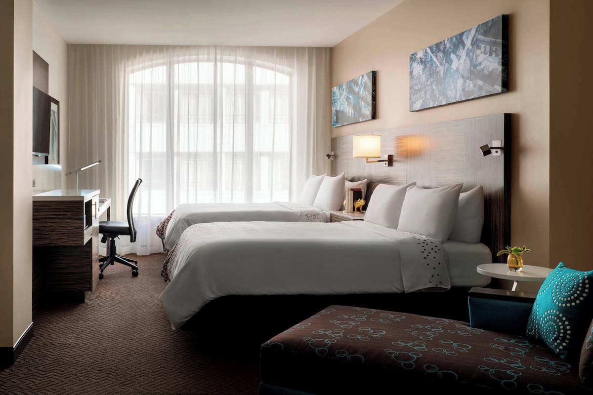 A hotel room with two beds, a desk, a chair, and a small table with a lamp.