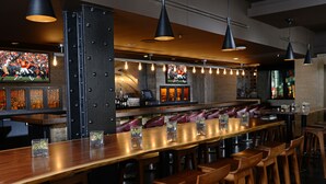 Una zona de bar con una barra de madera alargada, varios taburetes y luces colgantes.