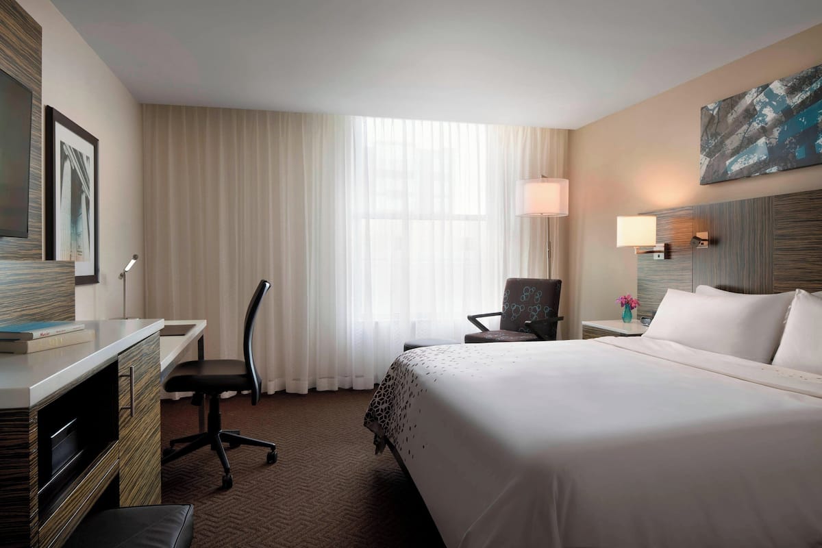 A hotel room with a large bed, a desk with a chair, a small table with a lamp, and a window with curtains.