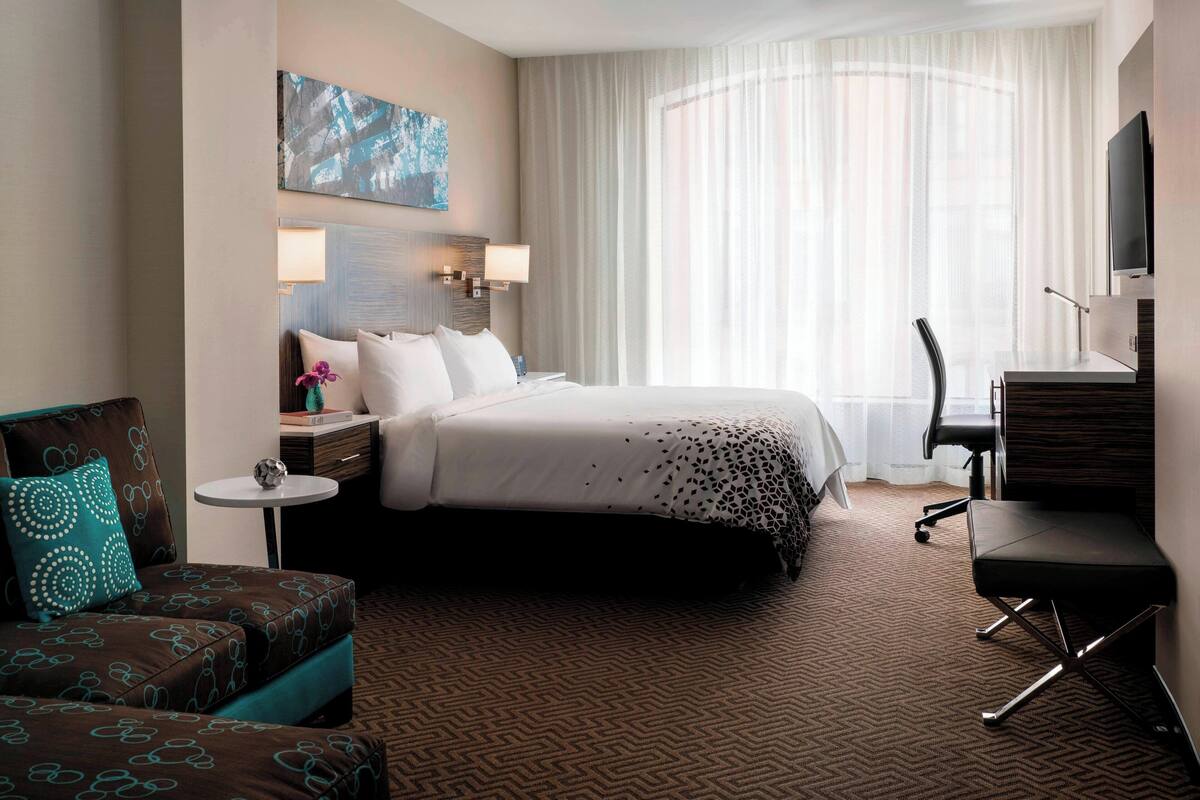 A hotel room with a bed, a desk, a chair, a small table with a lamp, and a painting on the wall.