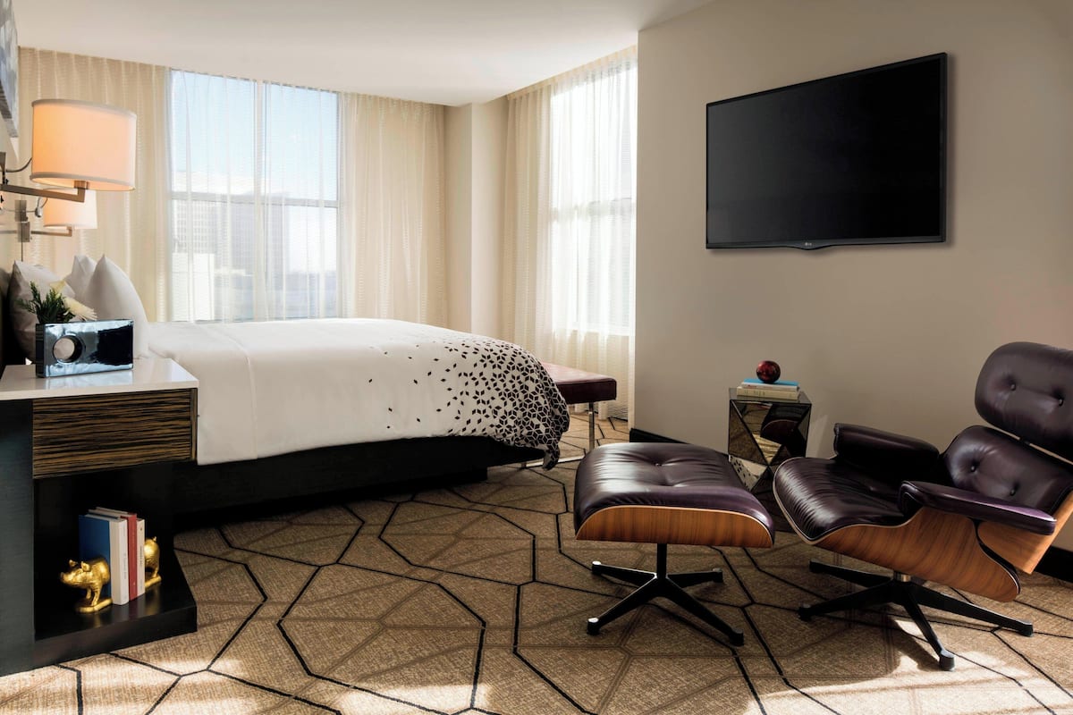 A hotel room with a large bed, a desk with a lamp, a leather armchair, and a flat-screen TV on the wall.