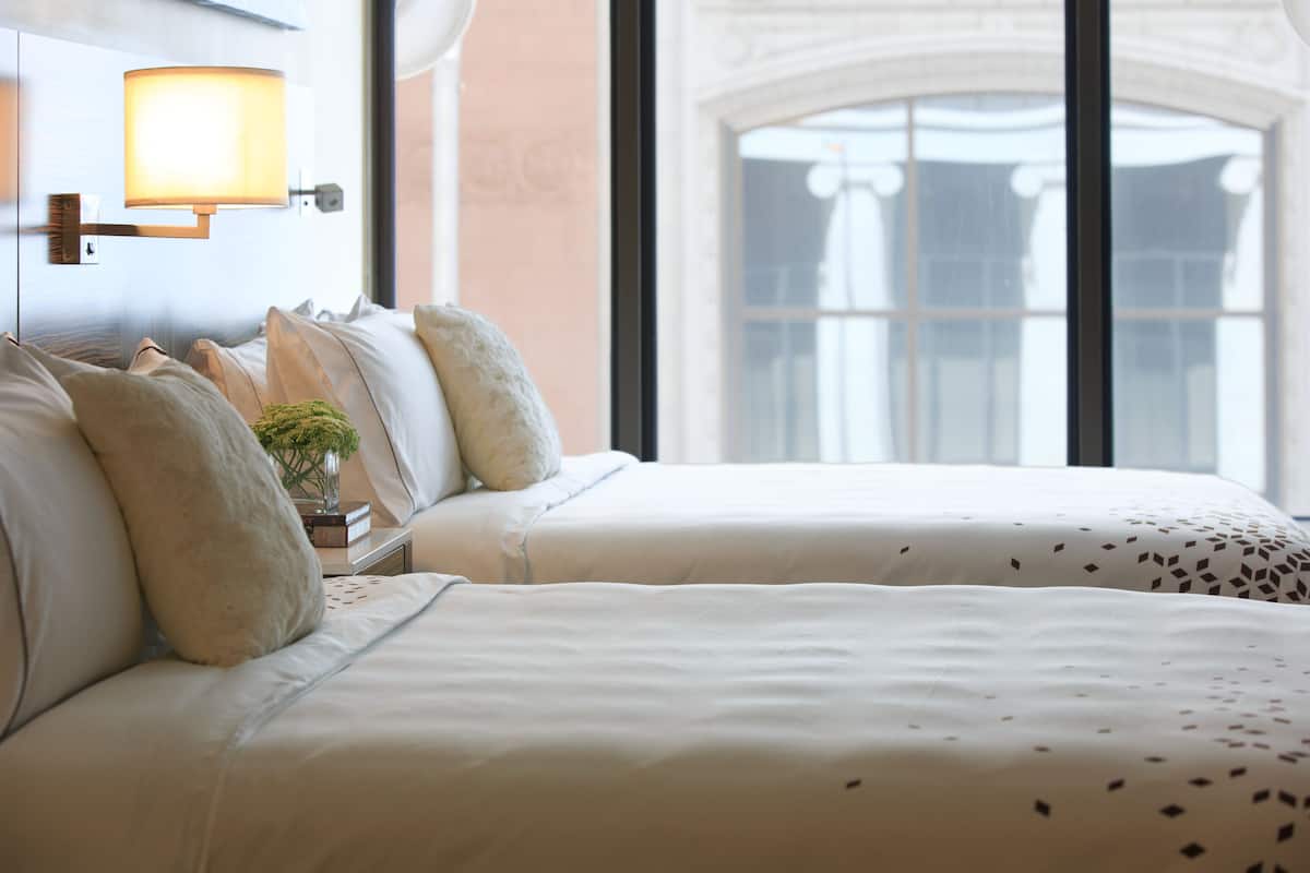 A hotel room with two beds, a nightstand with a lamp, and a view of a building through large windows.