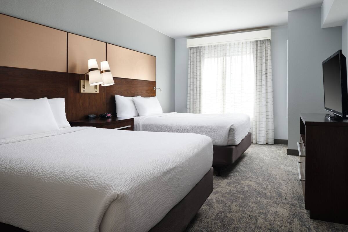 A hotel room with two beds, a wooden headboard, a flat-screen TV, and a large window with curtains.
