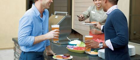 A group of people at an outdoor barbecue with a grill, plates of food, and drinks.