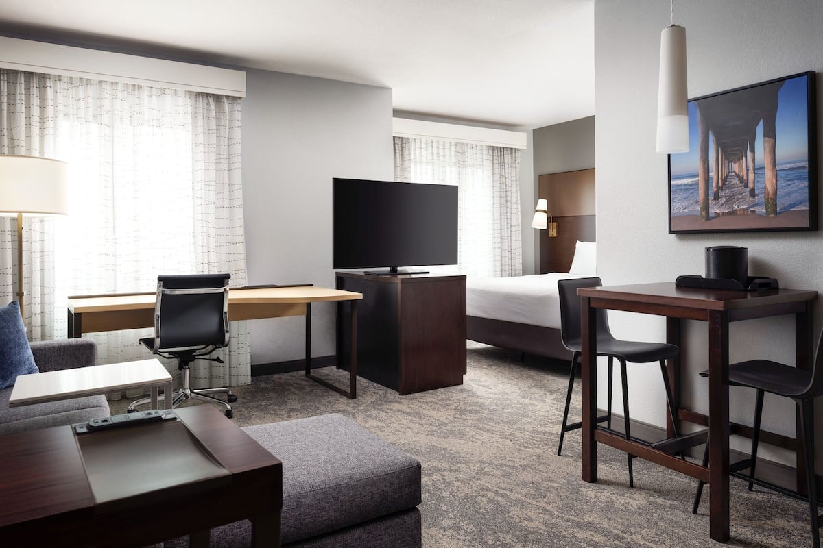 A hotel room with a bed, desk, chair, TV, and a painting of a pier.
