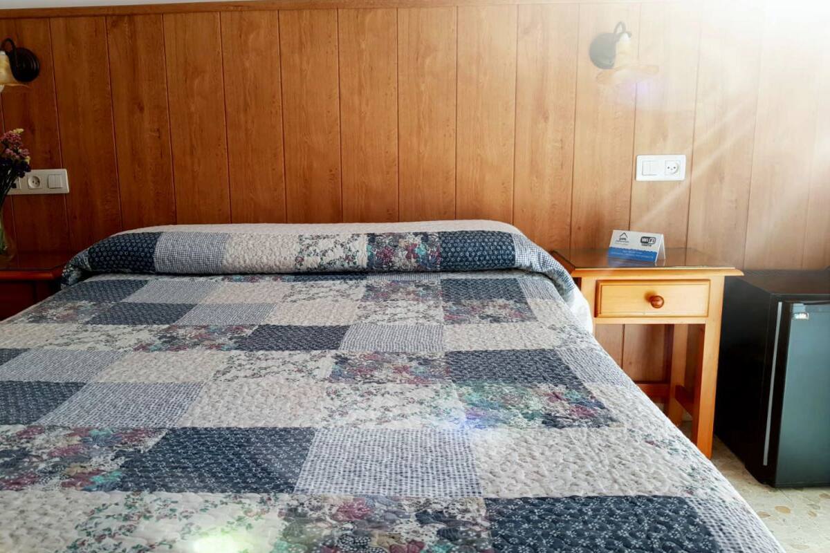 A room with a wooden paneled wall, a bed with a quilt, a nightstand with a lamp, and a small refrigerator.