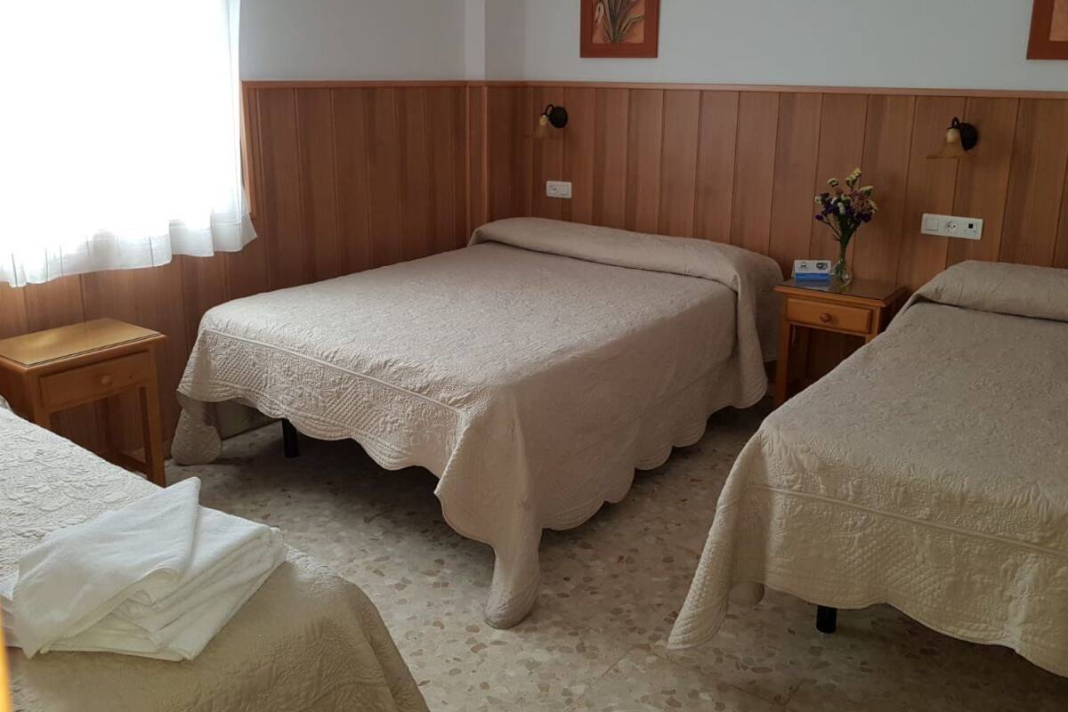 A hotel room with two beds, wooden paneling on the walls, a bedside table with a lamp, and a window with sheer curtains.