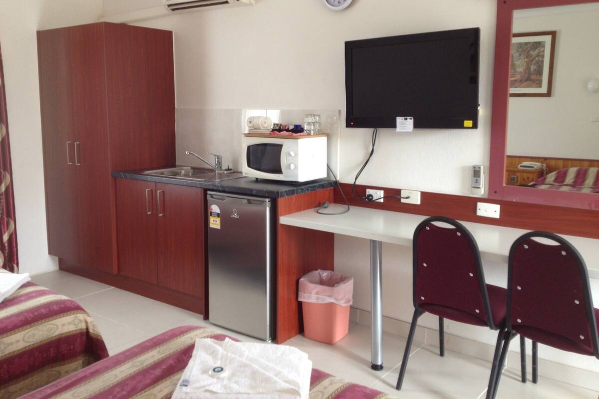 Cozinha compacta com micro-ondas, pia e um pequeno frigorífico. Há um relógio na parede e uma televisão fixada acima do balcão.