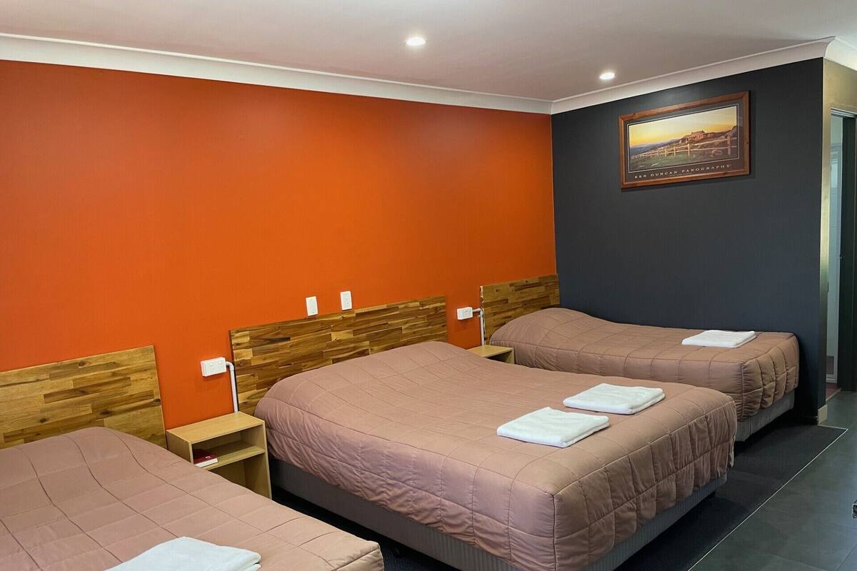 Quarto de hotel com duas camas, cada uma com cabeceira de madeira e cobertura rosa. Há uma mesinha de cabeceira com uma luminária e uma tomada na parede.