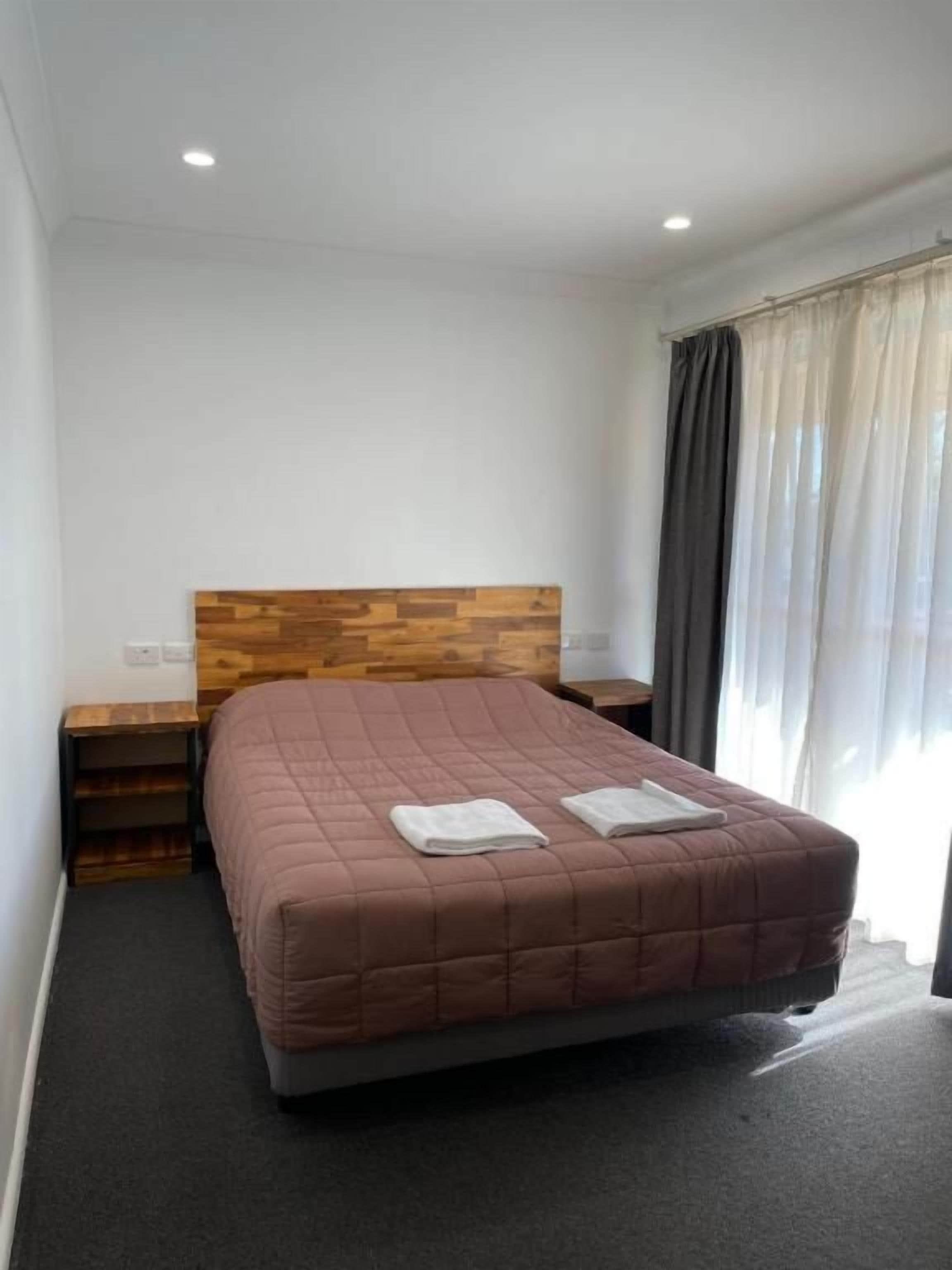 Una camera da letto con una testiera in legno, un letto con copriletto marrone, due comodini e una finestra con tende.