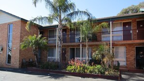 A two-story motel with a brick facade, balconies, and a small garden area with plants and a palm tree. - Homestead Motel (Dubbo)