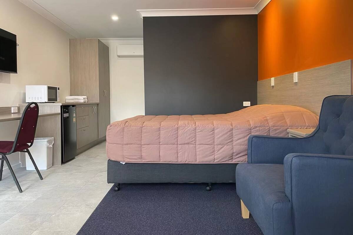 Quarto de hotel compacto com cama, escrivaninha, cadeira e micro-ondas.