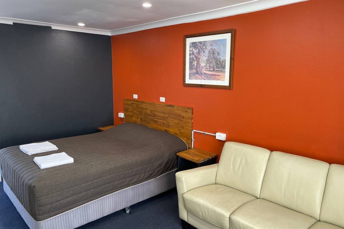 Quarto de hotel com cama, cabeceira de madeira, sofá bege e quadro na parede.