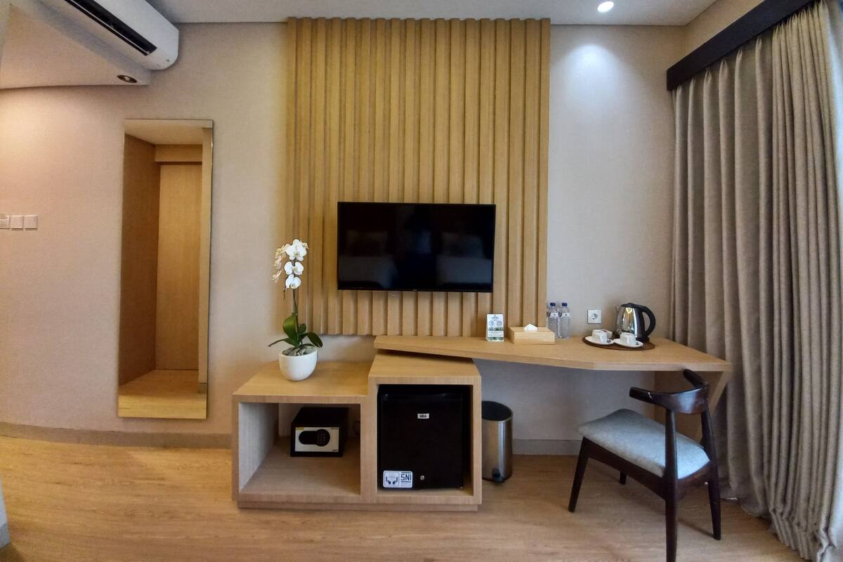 A modern hotel room with a flat-screen TV mounted on a wooden panel, a small seating area with a chair, a desk with a coffee maker, and a trash bin.