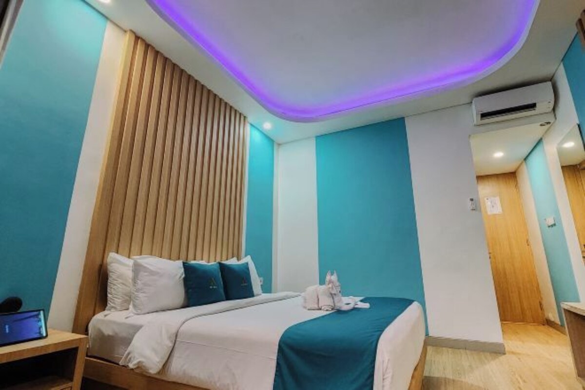 A modern hotel room with a bed, wooden headboard, teal walls, and a ceiling with purple lighting.