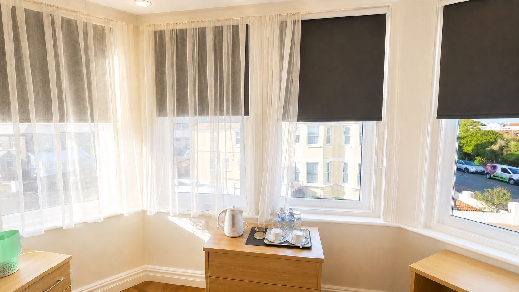 A room with a corner window, a wooden nightstand, a kettle, and a tray with cups and water bottles.