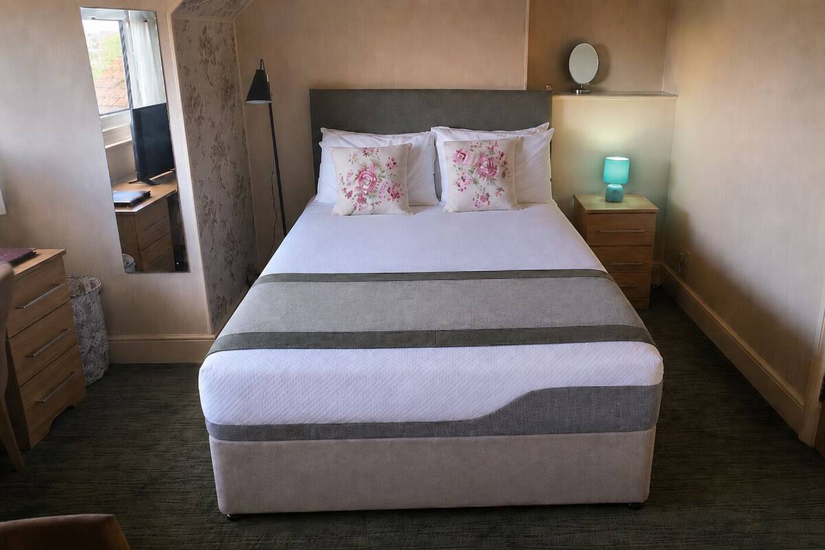A neatly made bed with pillows, a bedside lamp, a small table, and a mirror on the wall.