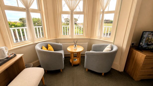 A room with two grey armchairs, a small round table with a candle, a bench, and a view of the sea through large windows. - Smartsway Hotel & Wellness (Eastbourne)