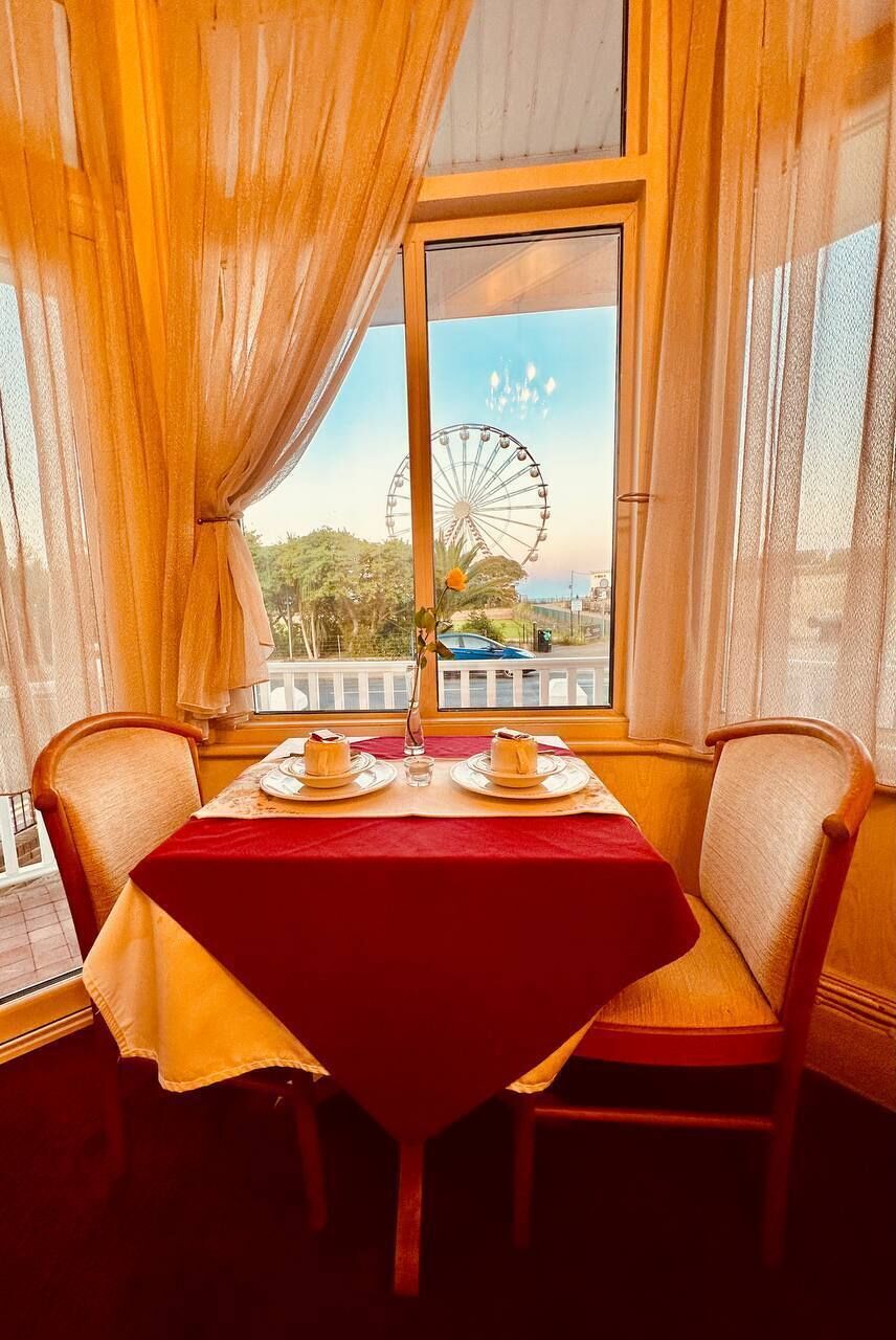A dining table with a red tablecloth, two chairs, and a view of a Ferris wheel through a window.