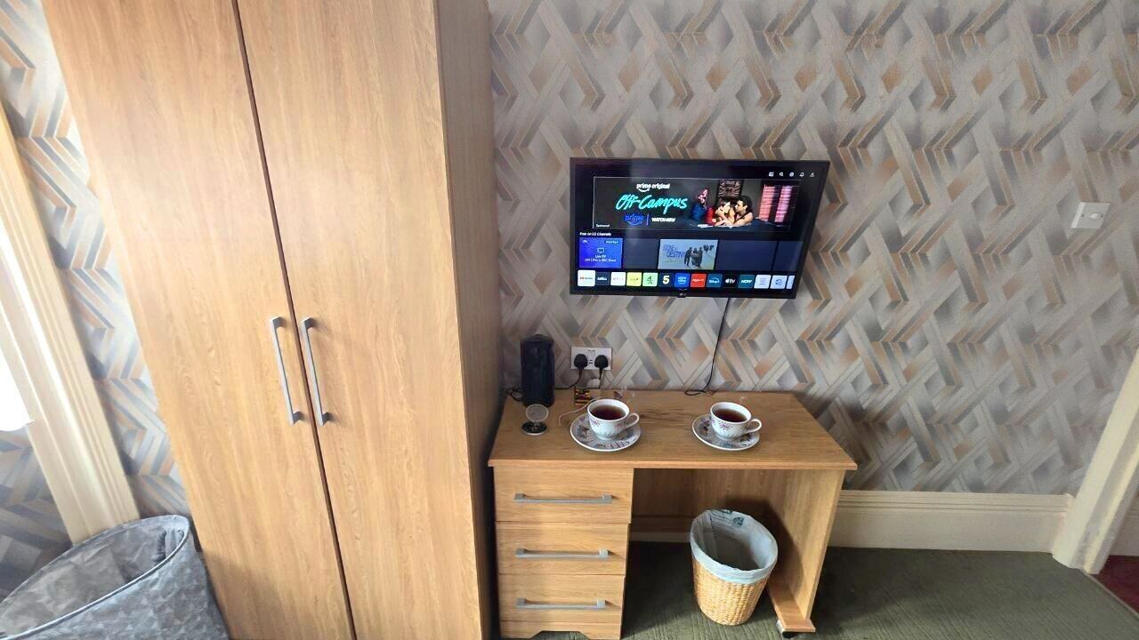 A room with a wooden wardrobe, a small wooden desk with two cups of coffee, a TV mounted on the wall, and a patterned wallpaper.