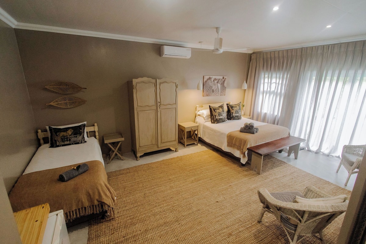 A hotel room with two beds, a wardrobe, a small table, a chair, and a large window with curtains.