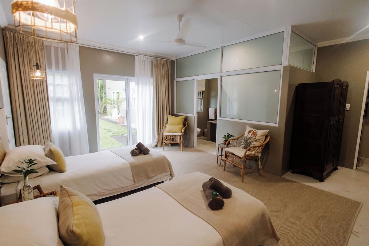 A hotel room with two beds, a sitting area with a chair and a small table, a large window, and a ceiling fan.