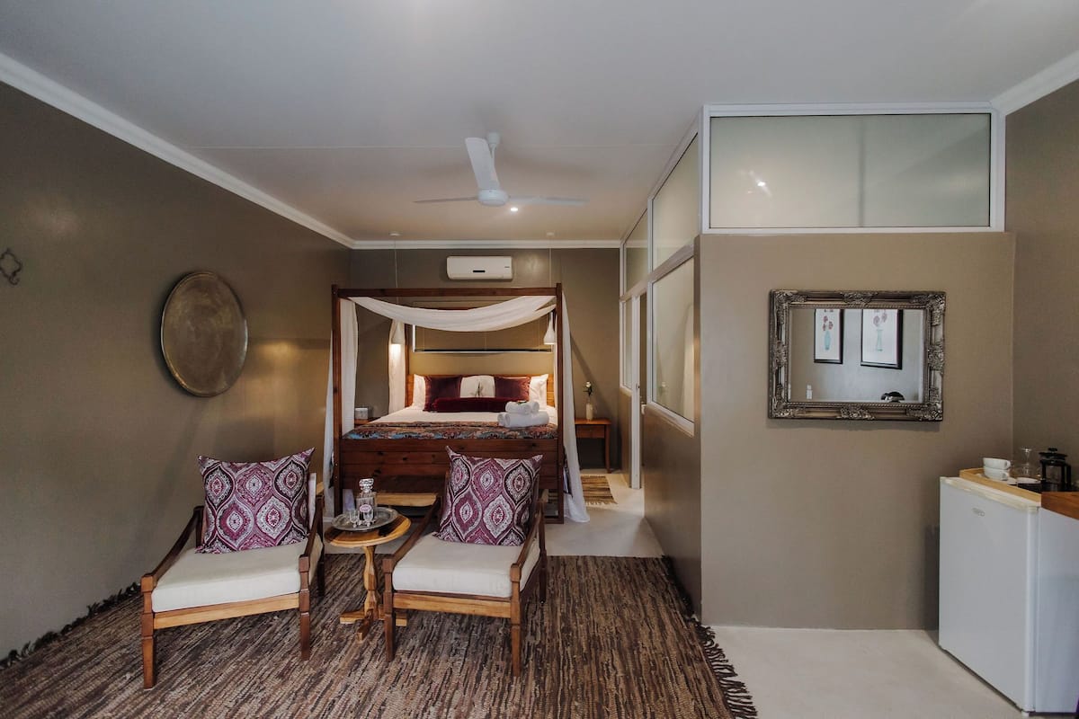 A hotel room with a bed, two chairs, a small table, a refrigerator, a ceiling fan, and a mirror on the wall.