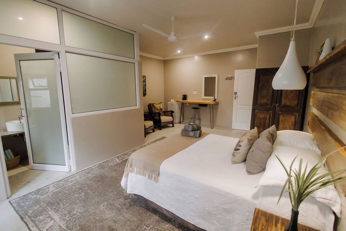 A modern bedroom with a large bed, a desk, a chair, and a bathroom with a glass door.