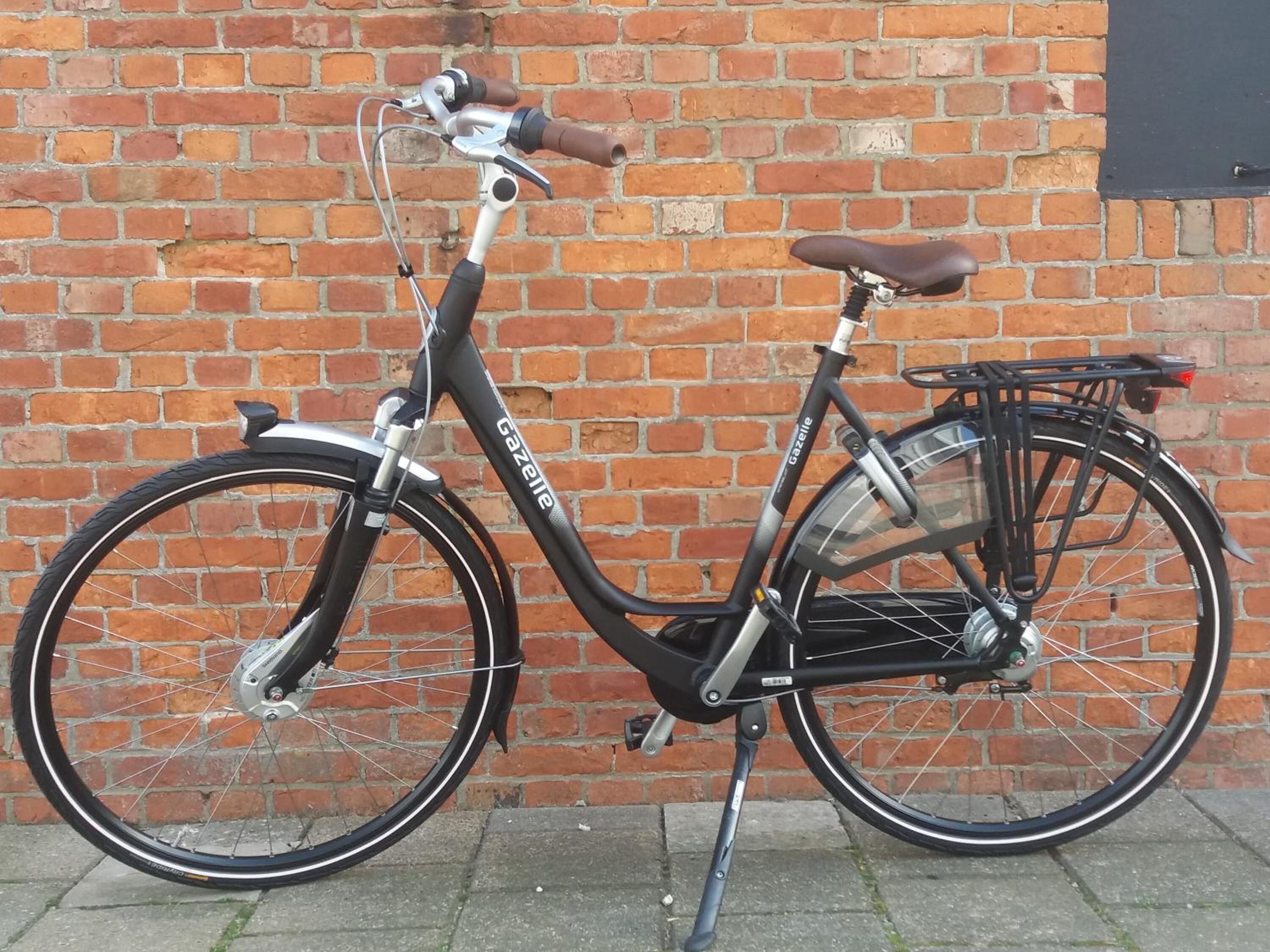 A stationary Gazelle electric bicycle with a brown seat and a rear rack, parked against a brick wall.