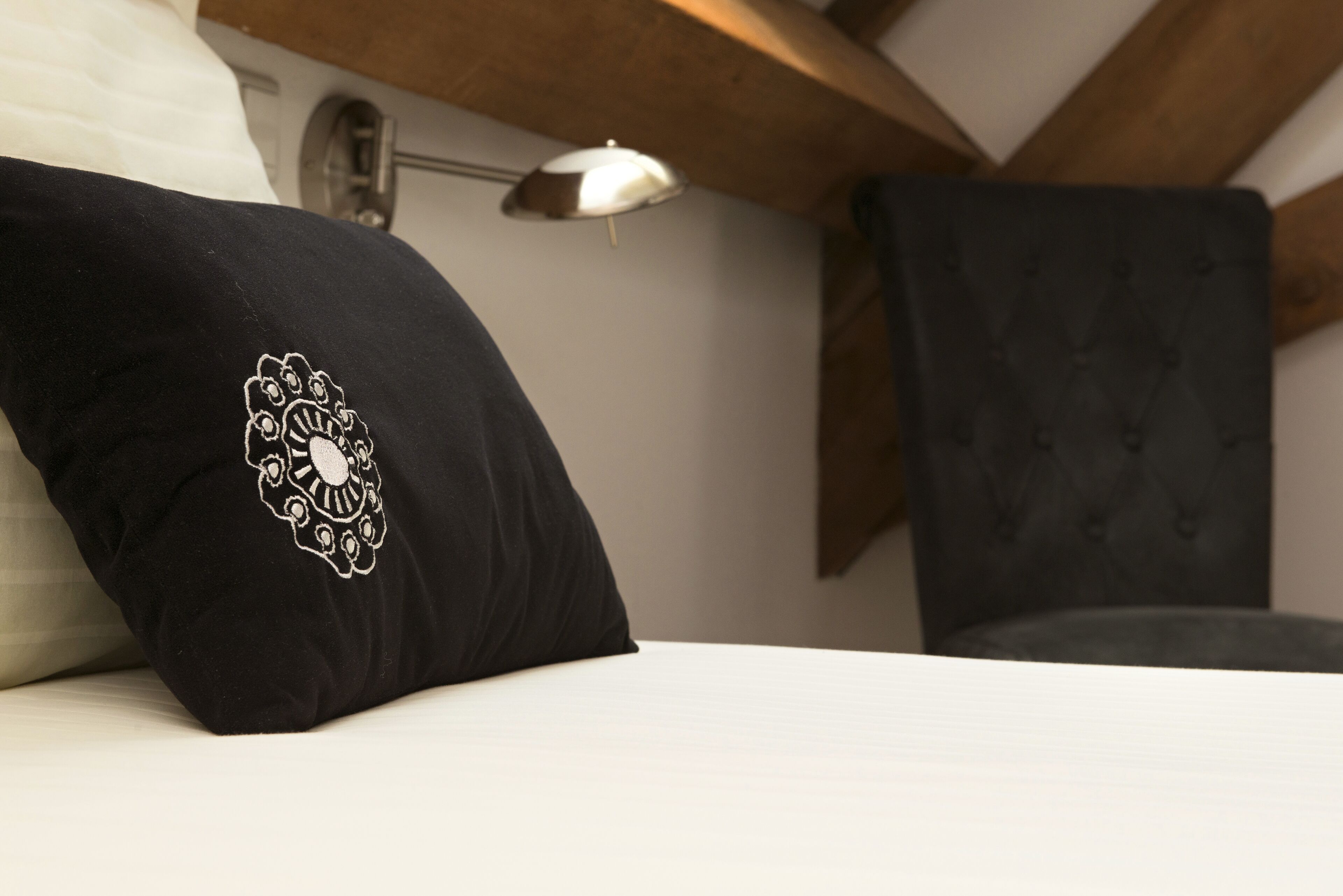 Una cama con un almohadón negro decorado con un diseño blanco bordado, una lámpara de noche y una butaca tapizada en tonos oscuros.