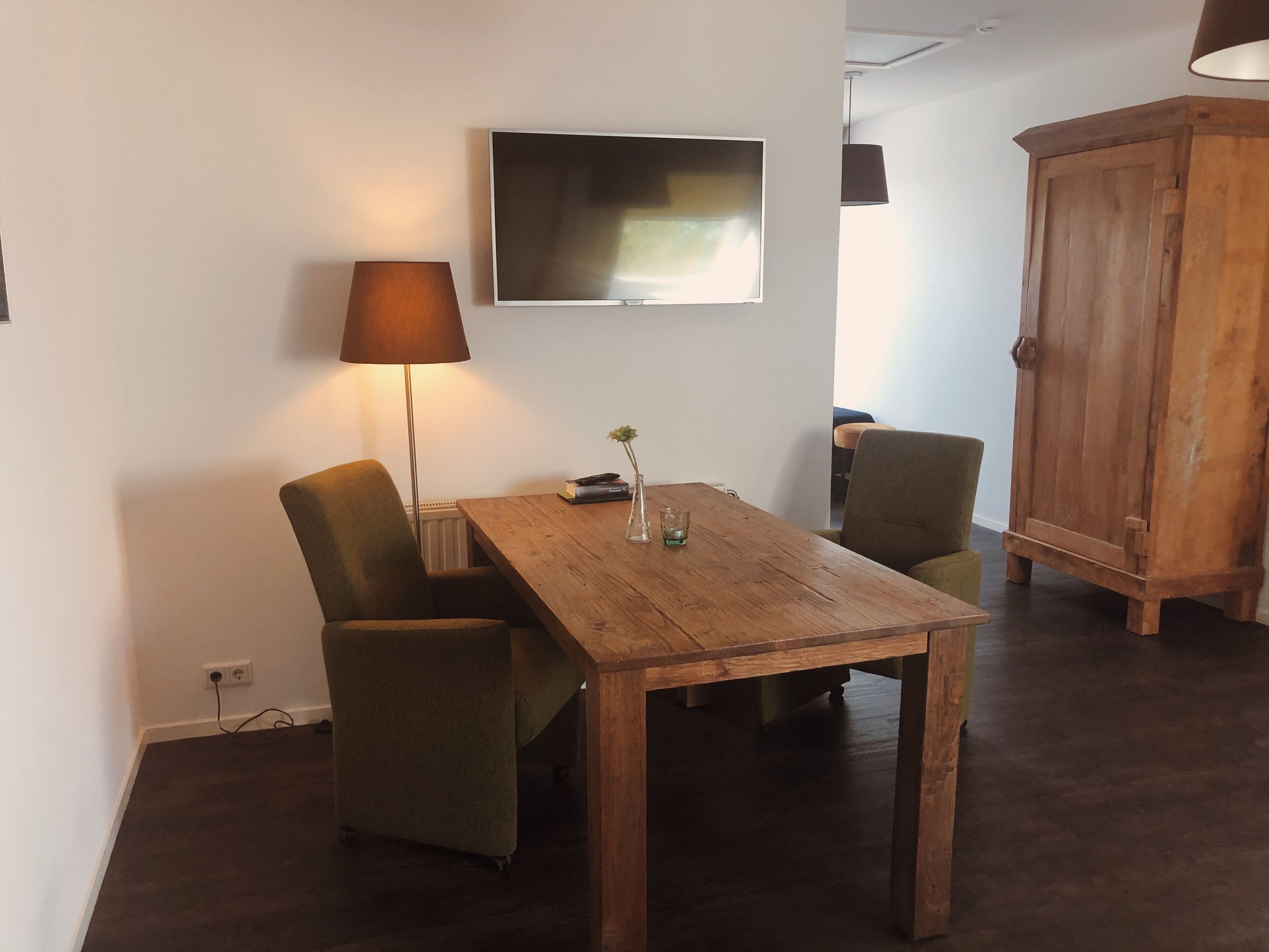 A wooden table with two chairs, a floor lamp, a television on the wall, and a wooden cabinet.