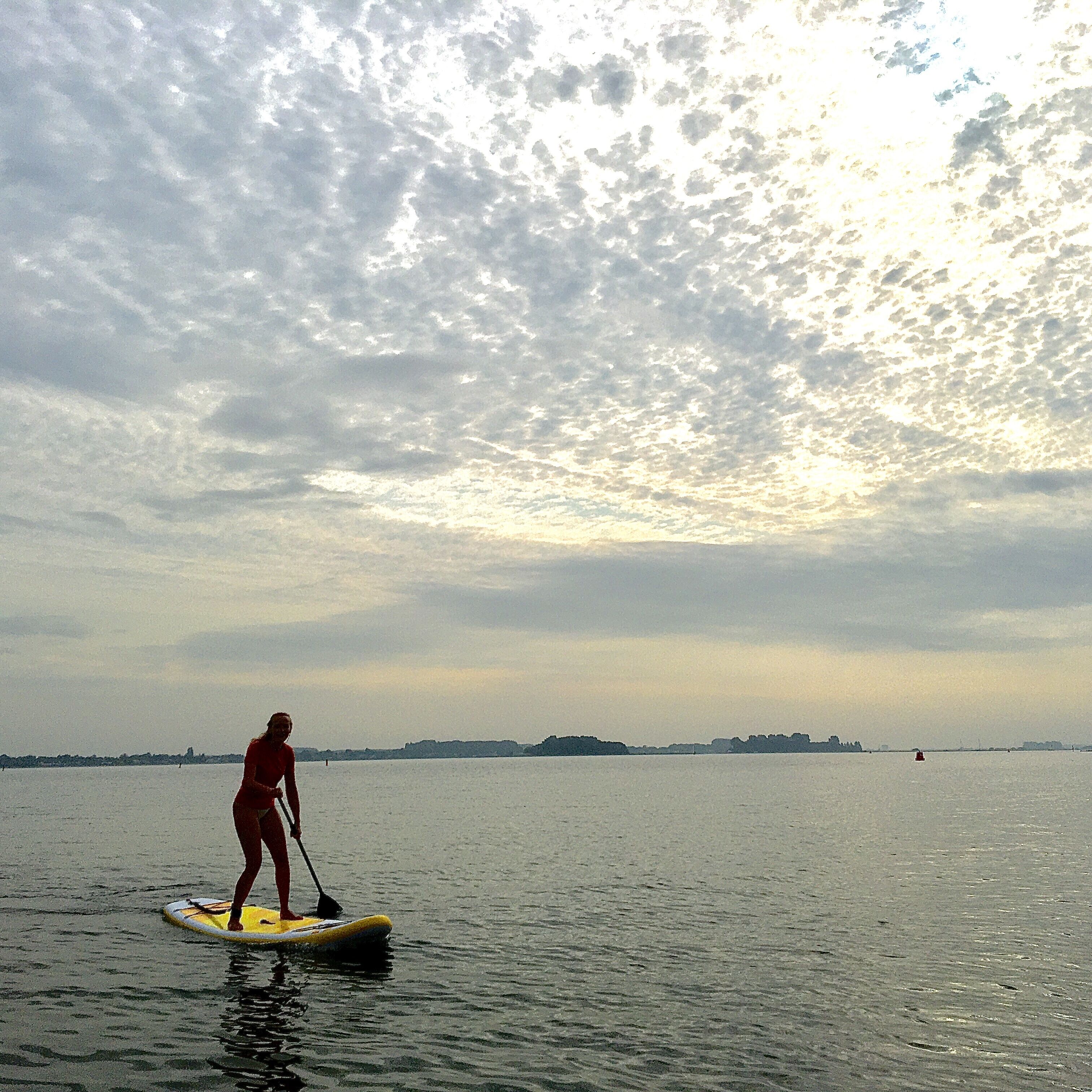 Une personne fait de la planche à pagaie sur une étendue d’eau calme, avec une côte lointaine et un ciel nuageux.