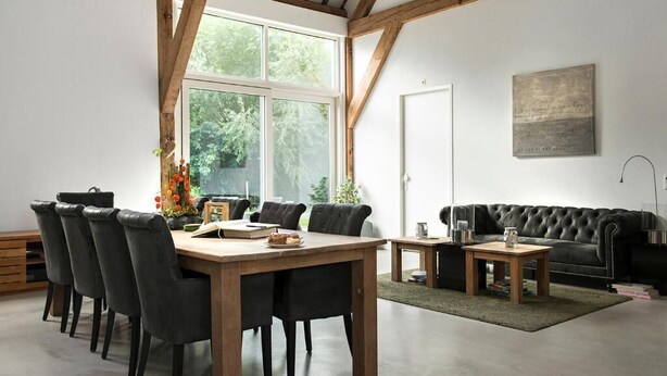 A spacious room with a wooden dining table and chairs, a black leather sofa, and a large window providing natural light. - Hoevehotel Hof Christina (Vrouwenpolder)