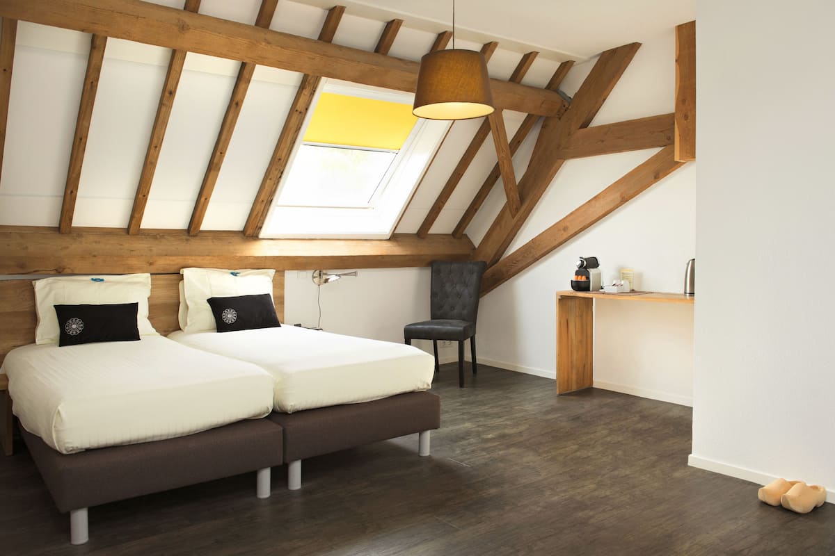 A bedroom with a sloped ceiling, wooden beams, a skylight, a bed with two pillows, a chair, and a small desk.