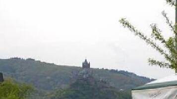 Ein Außenbereich mit Tischen und Stühlen, ein überdachter Bereich und ein Blick auf einen Hügel mit einer festungsähnlichen Struktur im Hintergrund.