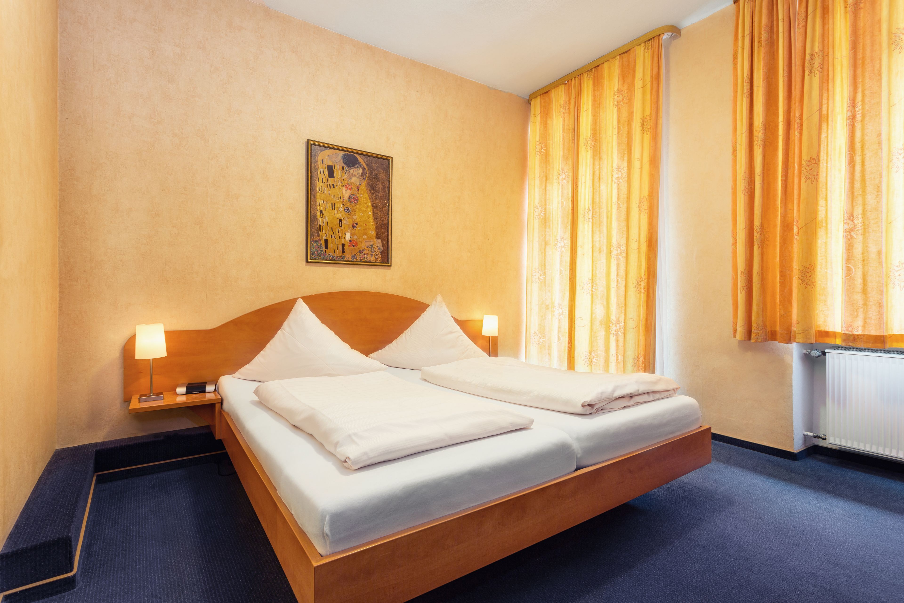 Ein Hotelzimmer mit einem hölzernen Bett, zwei Nachttischen mit Lampen, einem Bild an der Wand und einem Fenster mit Vorhängen.