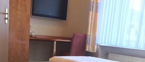 A hotel room with a bed, a television mounted on the wall, a window with curtains, and a wooden wardrobe.