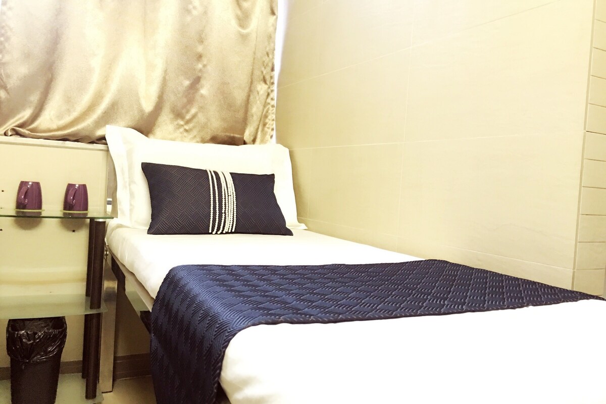 A small, neatly made bed with a dark blue quilt and a white pillow with a decorative string. There is a small table with a glass top and two purple cups beside the bed.