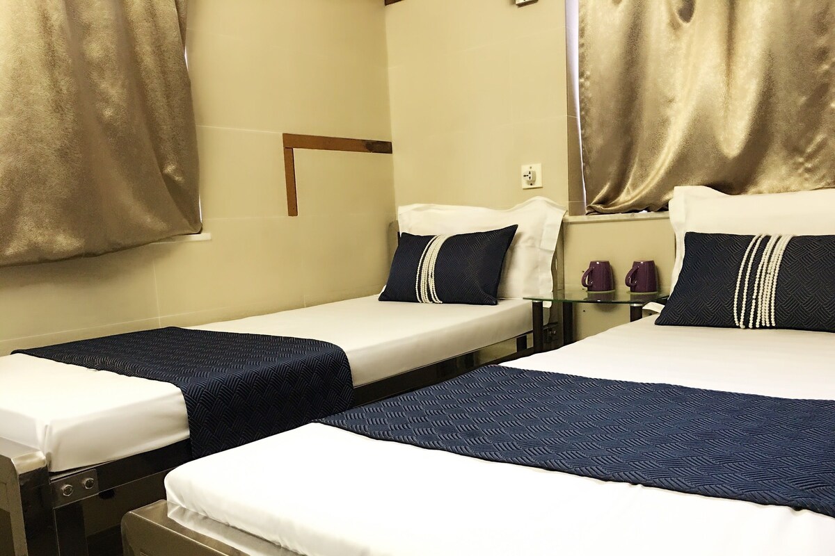 A hotel room with two single beds, each with a white mattress and navy blue bedspread. There are two bedside tables with lamps, a small table with a purple mug, and a wall-mounted light switch.