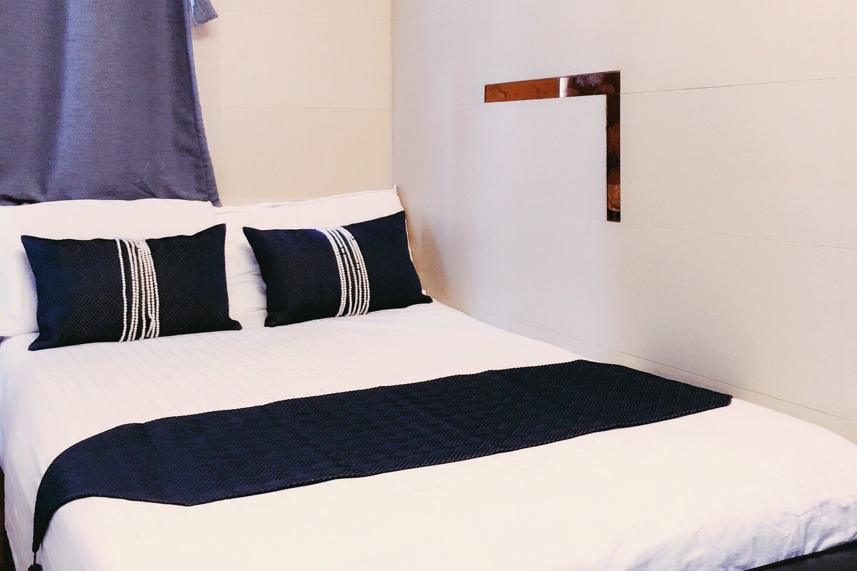 A neatly made bed with a white and dark blue bedspread, two pillows, and a blue curtain.