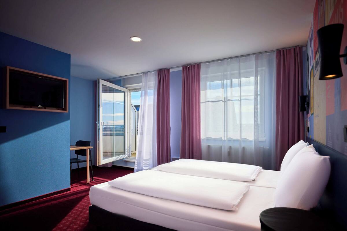 Habitación de hotel con cama, escritorio y silla. Se ve una ventana con cortinas y vistas al exterior.
