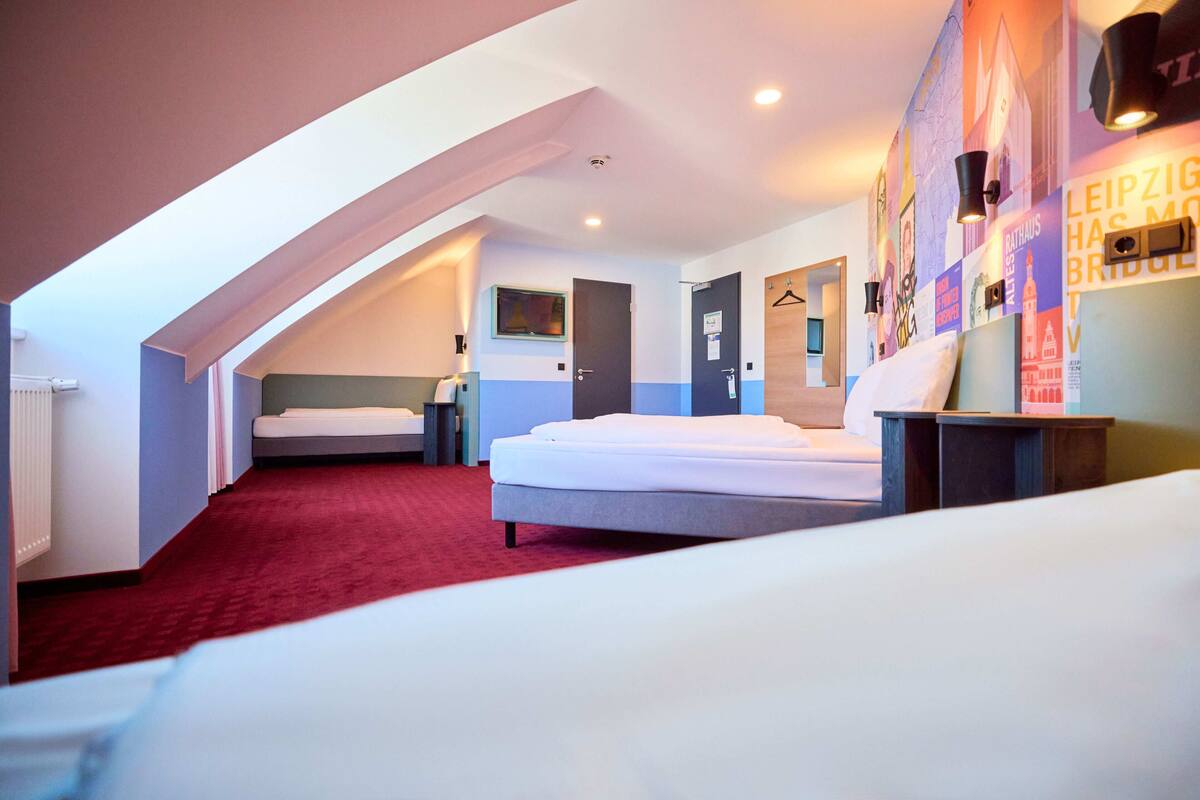 Habitación de hotel con techo inclinado, alfombra roja, cama, mesita de noche, armario y una pared con carteles.