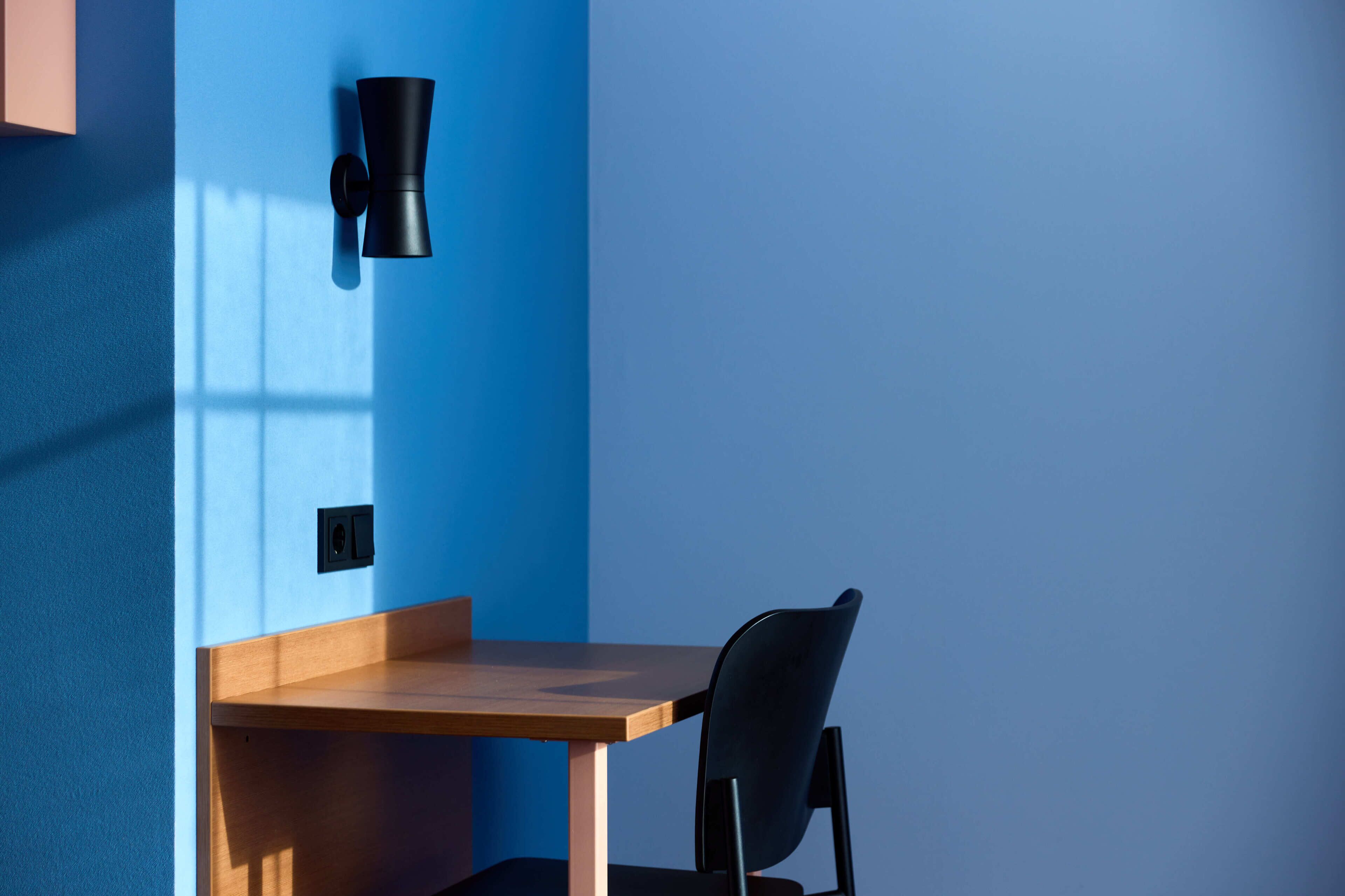 Et værelse med et træbord, en sort stol, en sort vægmonteret lampe og et vindue der kaster skygger.