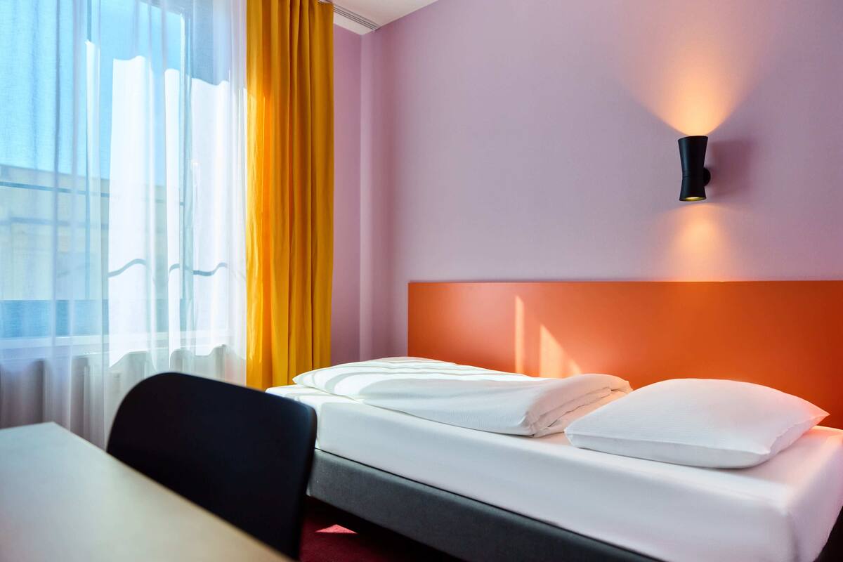 Habitación de hotel con cama, una silla, una mesa, ventana con cortinas y una lámpara de pared.