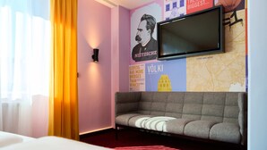 Une chambre d’hôtel avec un canapé gris, un téléviseur à écran plat fixé au mur et une grande affiche représentant un paysage urbain.