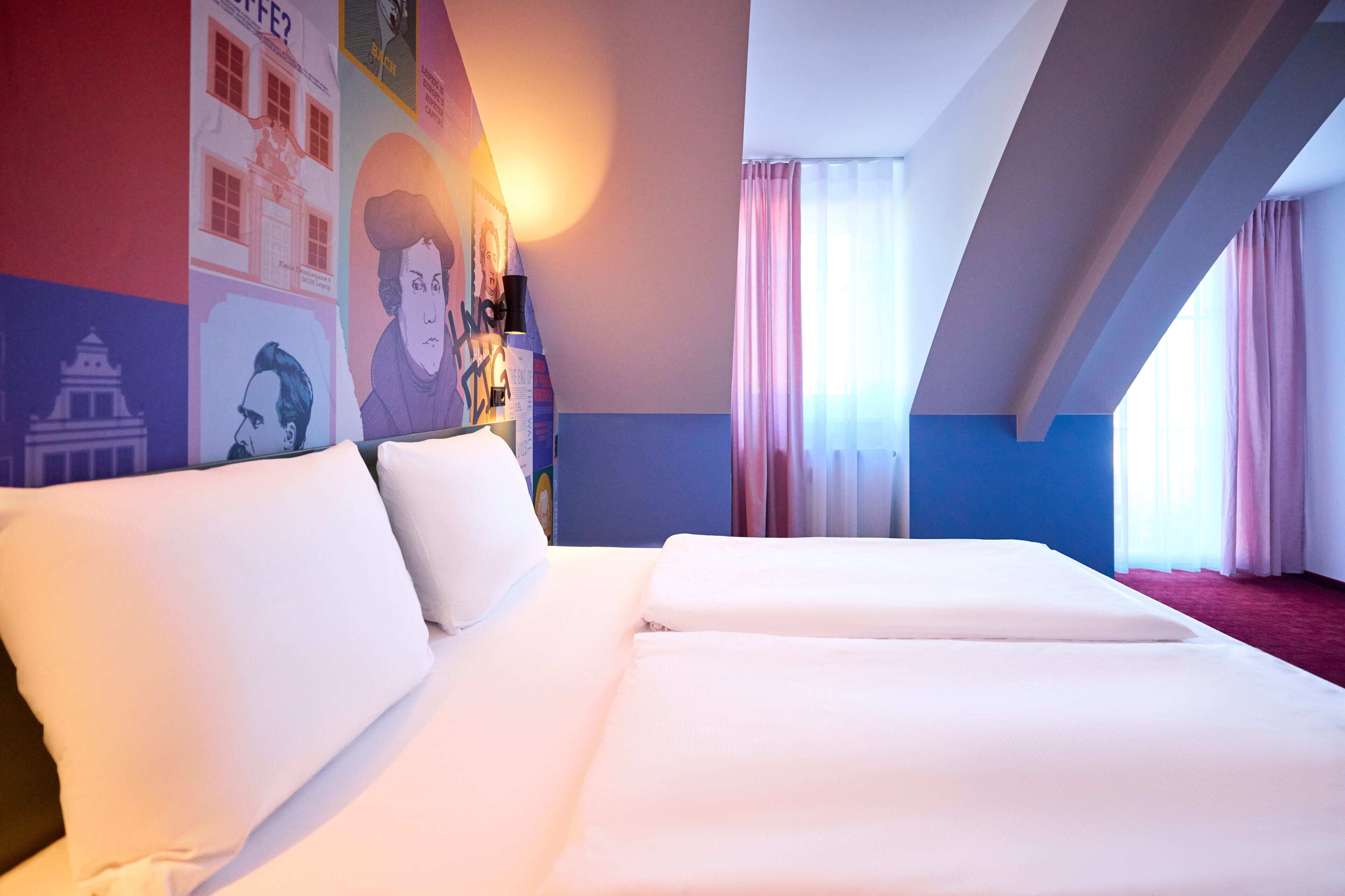 Een hotelkamer met twee bedden, een groot raam met gordijnen en een muur versierd met posters.