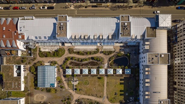Vue aérienne d’un ensemble de bâtiments dotés de toits multiples, d’une cour centrale agrémentée de verdure et d’un espace de stationnement.