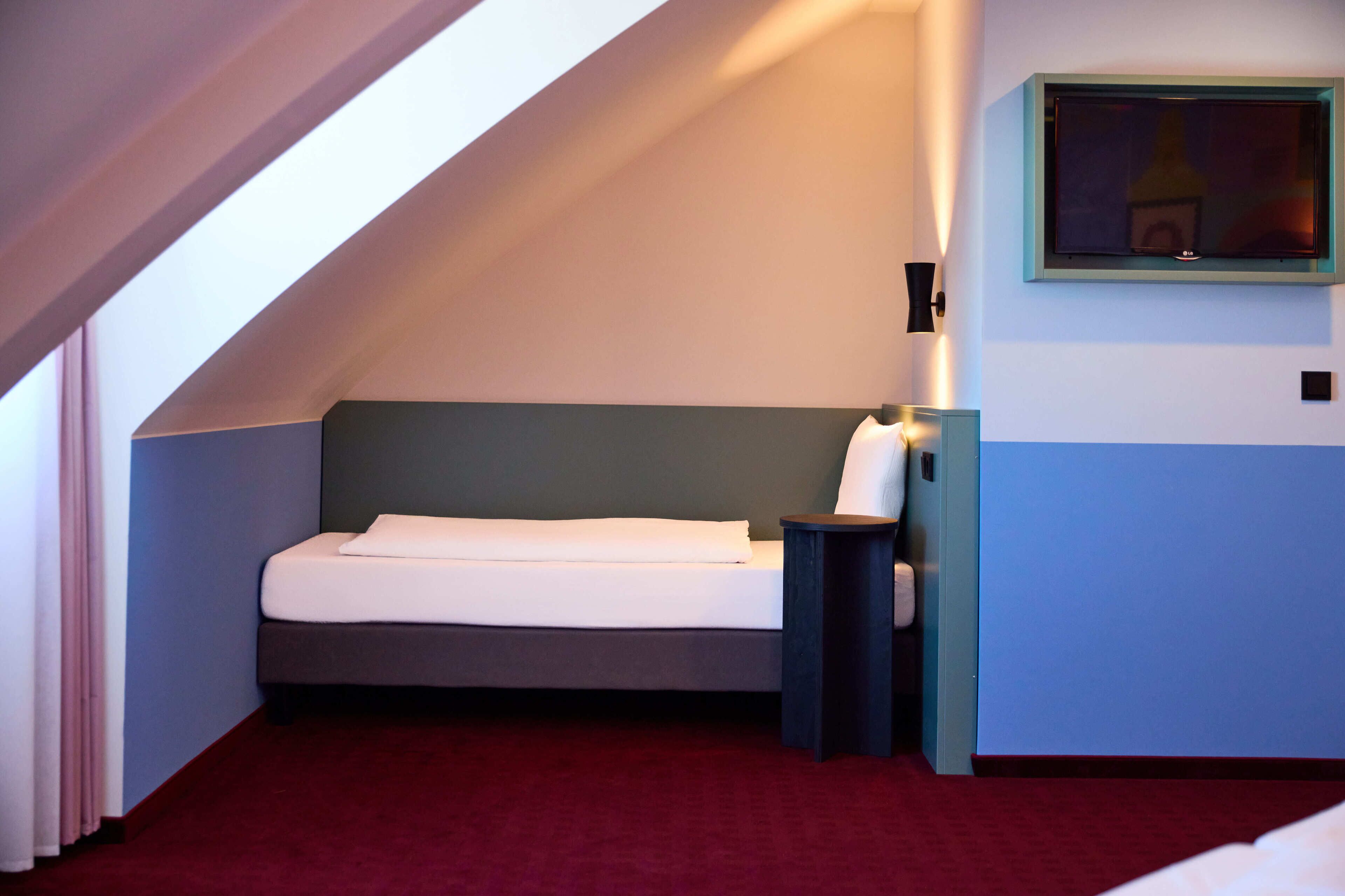 Een klein, verzorgd bed in een kamer met een schuine kap, een nachtkastje met een lamp en een aan de muur bevestigde televisie.